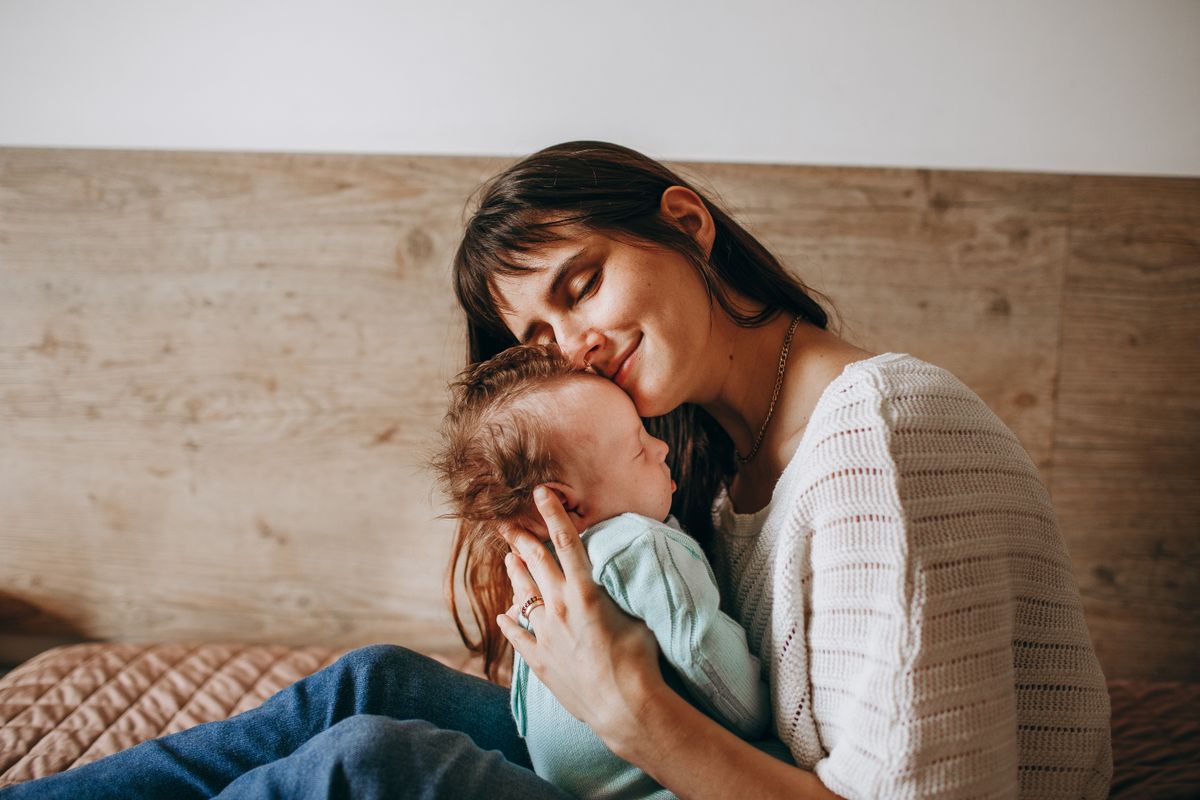 A mother sitting on a bed, eyes closed, holding her sleeping newborn against her cheek