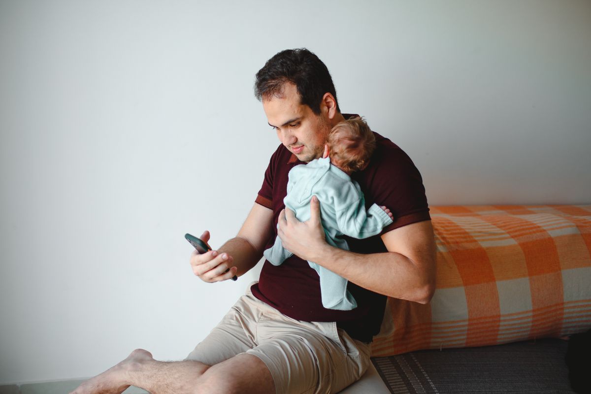 A father sitting on a sofa holding his sleeping newborn against his shoulder while checking a baby sleep app on his phone
