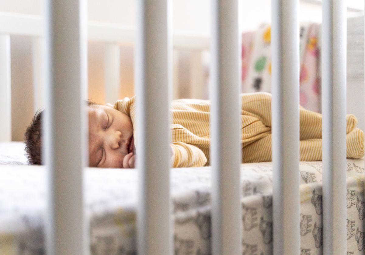 A newborn in a striped sleepsuit napping on their back in a white crib, seen through the bars
