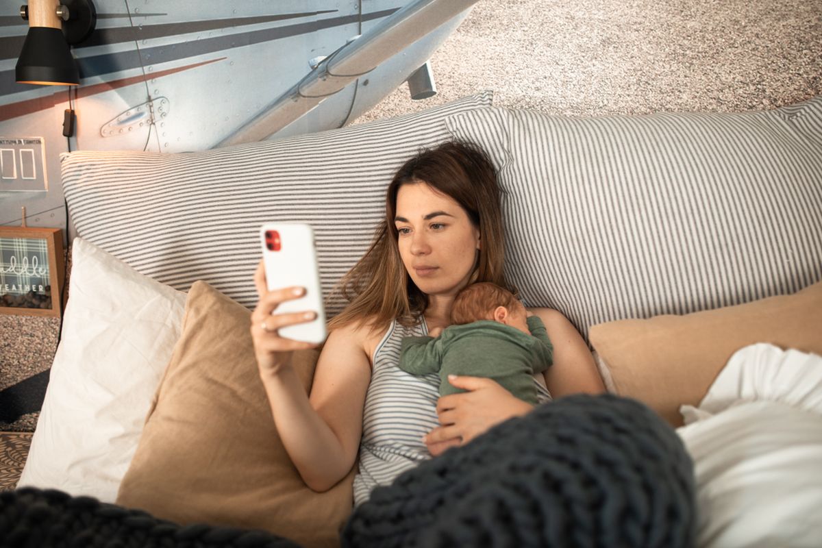 A mother lying in bed with her newborn asleep on her chest, checking a baby sleep app on her phone