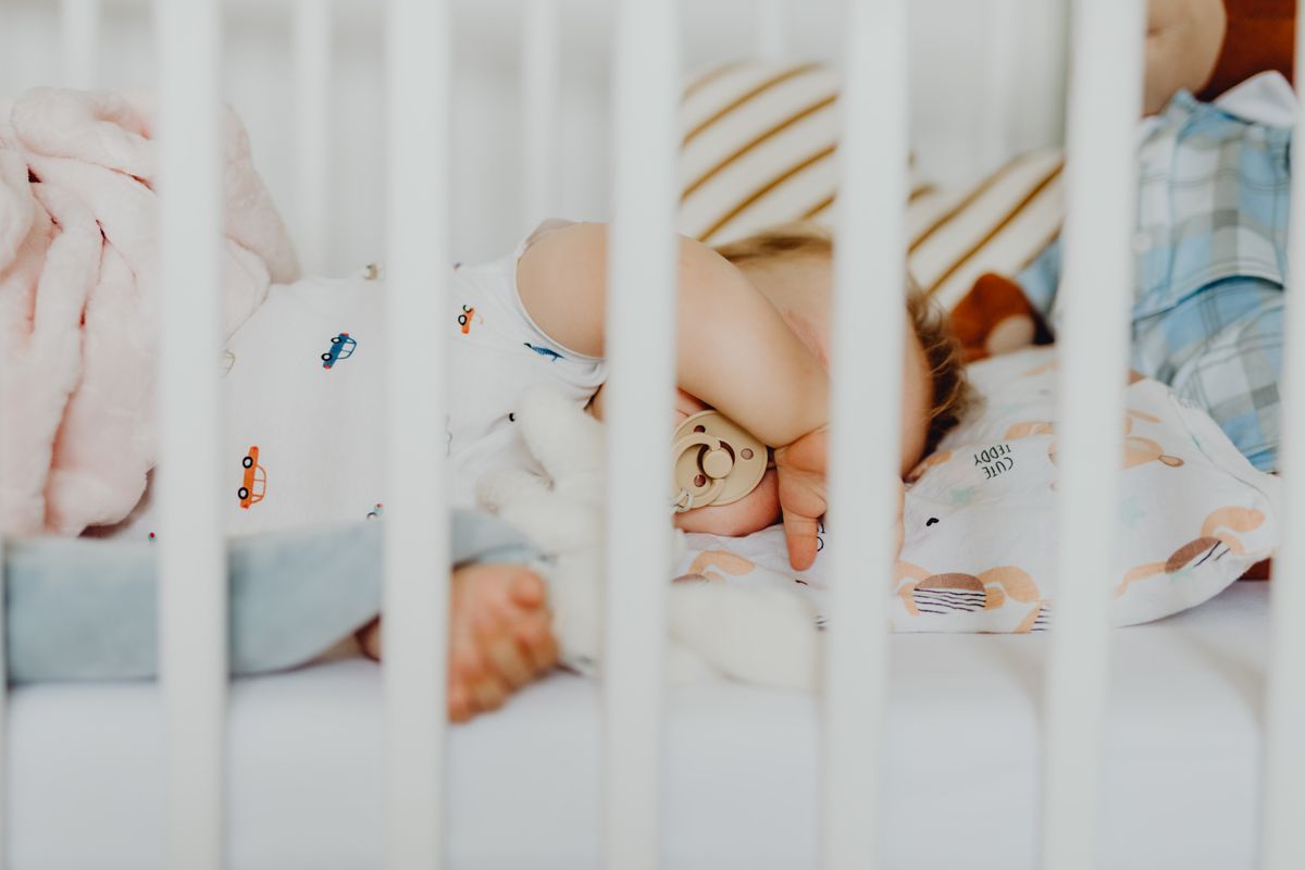 A calm baby lying awake in a crib in a dimly lit nursery while a parent stands nearby
